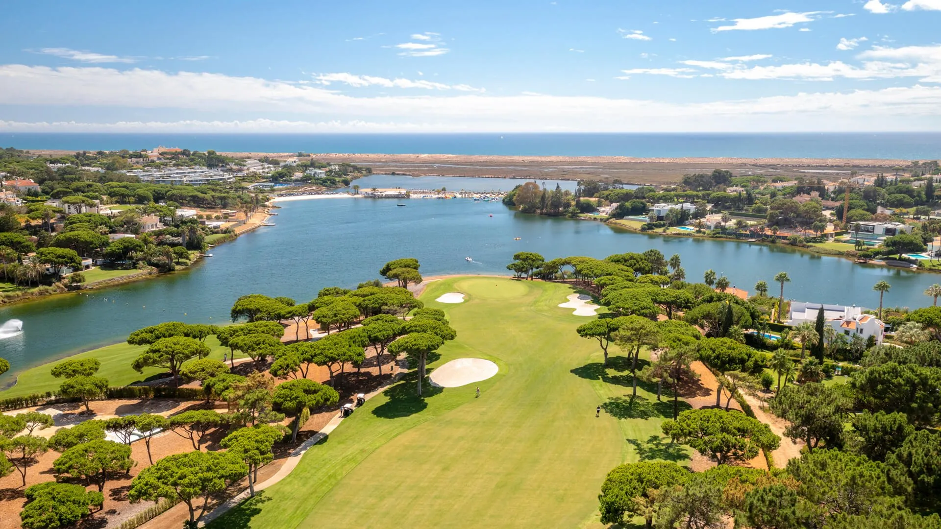 Stunning aerial view of an elite Algarve golf course next to a tranquil lake/lagoon, surrounded by luxury villas and green pine trees, with the Atlantic Ocean in the background.
