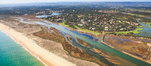 Wide aerial landscape view of the Algarve coastline, showing the sandy beach, the winding Ria Formosa lagoon, and luxury residential properties on the green mainland.